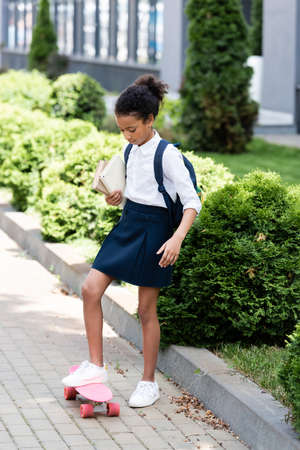 african american schoolgirl with backpack and books riding penny board outdoorsの写真素材