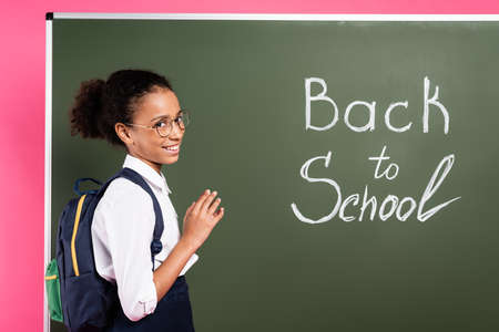 smiling african american schoolgirl in glasses near back to school inscription on green chalkboard on pink backgroundの写真素材