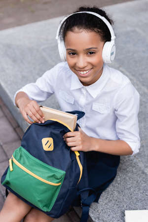 happy african american schoolgirl in headphones putting book in backpack