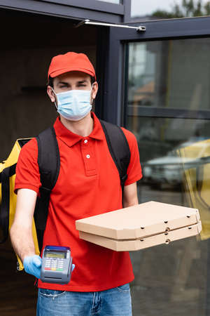 Courier in medical mask and latex glove holding payment terminal and pizza boxes on urban streetの写真素材