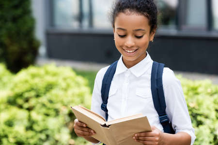 smiling african american schoolgirl reading book outdoorsの写真素材