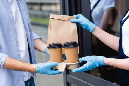 Cropped view of man receiving paper cups and package from waitress in latex gloves near cafeの写真素材