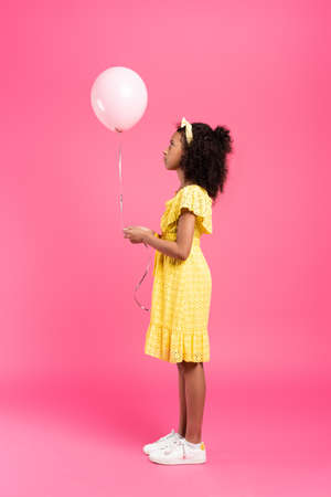 side view of curly african american child in yellow outfit with balloon on pink backgroundの写真素材
