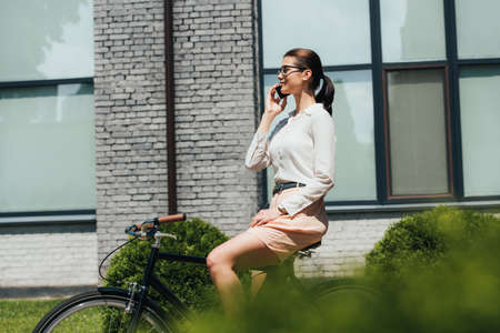 selective focus of businesswoman in glasses talking on smartphone and sitting on bicycleの写真素材