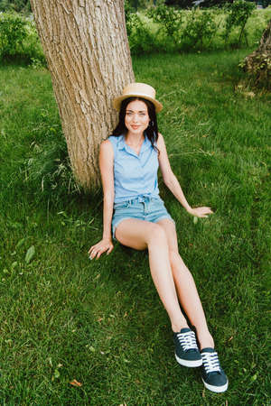 happy girl in straw hat sitting on green grass near tree trunk