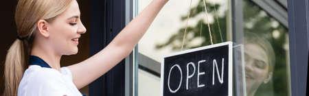 Panoramic shot of smiling waitress hanging signboard with open lettering on door of cafeの写真素材