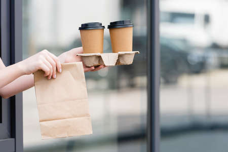 Cropped view of waitress holding paper bag and coffee to go near cafe on urban streetの写真素材