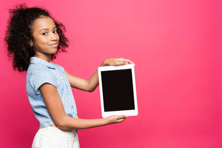 smiling cute curly african american kid presenting digital tablet isolated on pinkの写真素材
