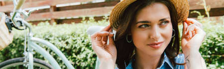 horizontal image of attractive woman touching straw hat near bicycleの写真素材