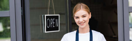 Panoramic shot of smiling waitress looking at camera near signboard with open lettering on door of cafeの写真素材