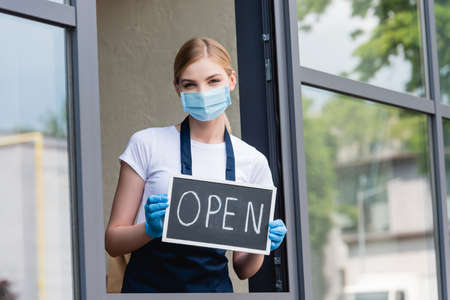 Waitress in latex gloves and medical mask holding signboard with open word near window of cafe