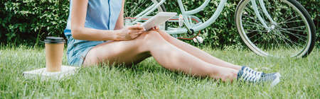 panoramic crop of girl holding digital tablet and sitting on grass near bicycle, paper cup and book
