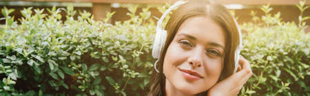 panoramic shot of cheerful young woman listening music in wireless headphones near bushの写真素材