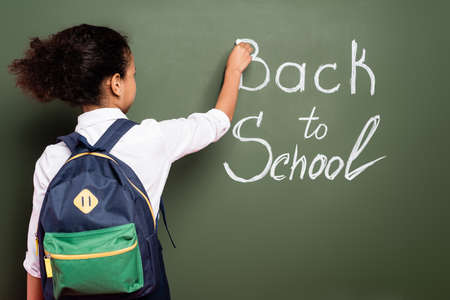 back view of african american schoolgirl with backpack writing back to school inscription on green chalkboardの写真素材