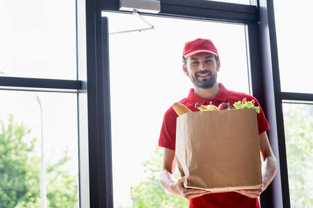 Smiling courier holding shopping bag with grocery and looking at cameraの写真素材