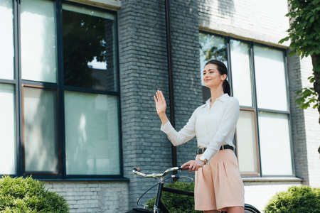 cheerful young businesswoman standing with bike and waving handの写真素材