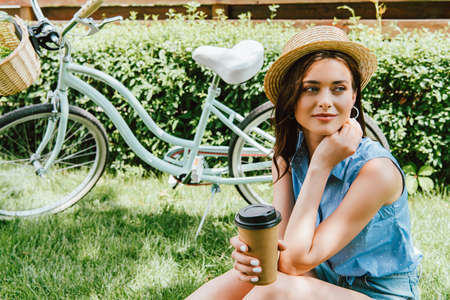girl in straw hat holding paper cup and sitting near bicycleの写真素材