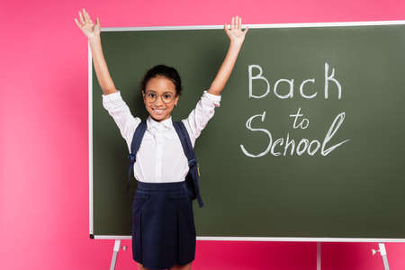 happy african american schoolgirl in glasses with hands in air near back to school inscription on green chalkboard on pink backgroundの写真素材