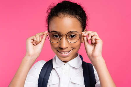 smiling african american schoolgirl adjusting glasses isolated on pinkの写真素材