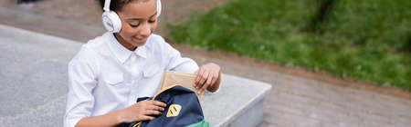happy african american schoolgirl in headphones putting book in backpack, panoramic shotの写真素材