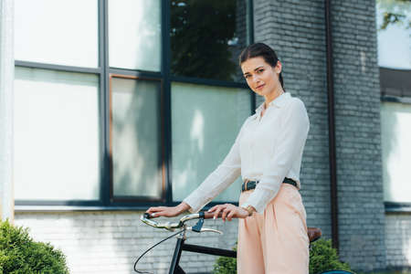 beautiful young businesswoman standing with bike near modern buildingの写真素材