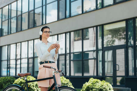 attractive businesswoman in glasses listening music in wireless headphones and using smartphone near bicycleの写真素材