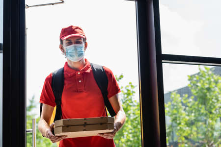 Courier in medical mask holding pizza boxes near open door of buildingの写真素材