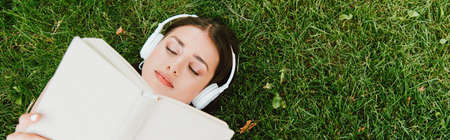 panoramic shot of girl in wireless headphones reading book and lying on green grass