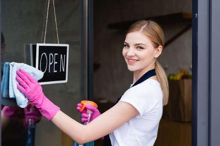 Side view of waitress smiling at camera while cleaning door of cafe near signboard with open letteringの写真素材