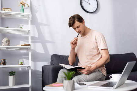 Selective focus of pensive freelancer holding notebook and pen near gadgets and coffee on coffee...