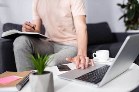 Cropped view of freelancer using laptop and writing on notebook near coffee and smartphone on coffee...