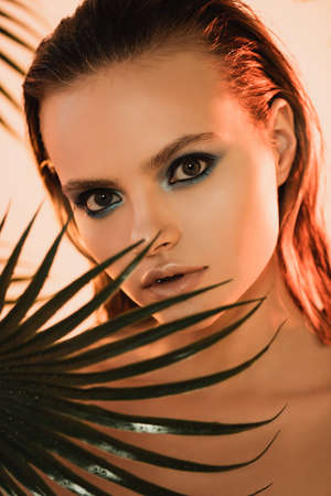 selective focus of beautiful woman with blue eye shadow looking at camera near palm leaf