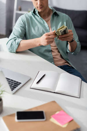 Cropped view of man holding cash near digital devices and notebooks on table at home, earning online conceptの写真素材