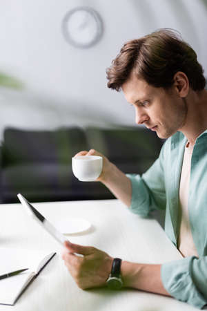 Selective focus of handsome man holding cup of coffee and using digital tablet at table, concept of earning onlineの写真素材