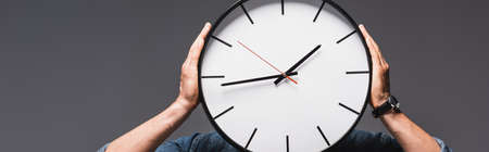 Panoramic crop of man holding clock near face isolated on gray, concept of time managementの写真素材