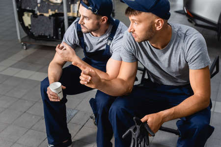 Auto mechanic sitting and holding paper cup near colleague pointing with finger at service station