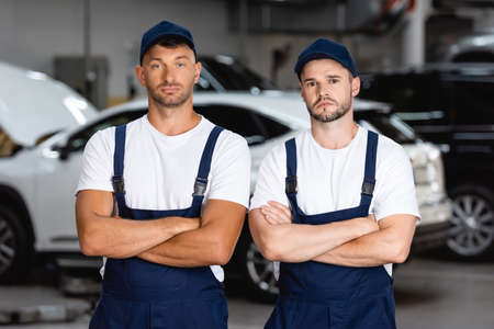 handsome mechanics in uniform and caps standing with crossed arms in car serviceの写真素材