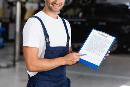 cropped view of happy mechanic in overalls and cap holding clipboard with contract lettering and pen near carsの写真素材