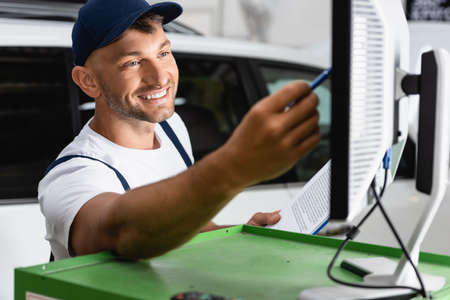 selective focus of happy mechanic in cap holding pen near computer monitor in workshop