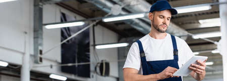 panoramic shot of mechanic in uniform and cap using digital tablet in workshop