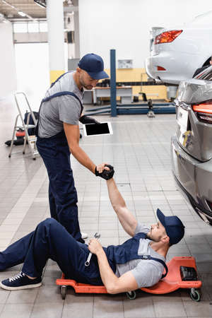 mechanic with digital tablet holding hands with coworker in cap lying near car