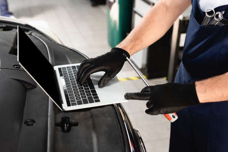 Cropped view of auto mechanic typing on laptop on car and holding screwdriver at service stationの写真素材