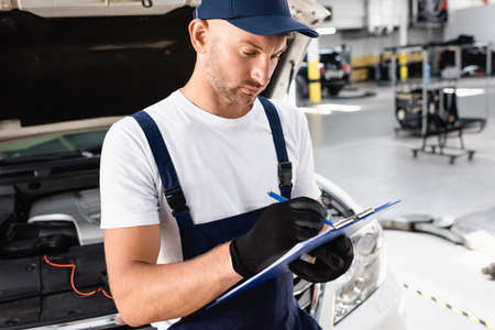 Auto mechanic writing on clipboard near open hood of car at service station
