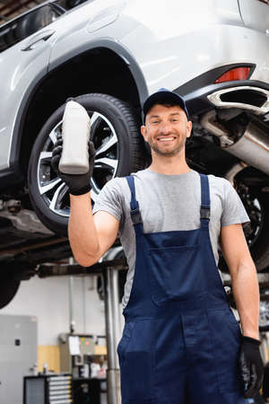 low angle view of cheerful mechanic in cap holding bottle with car oil