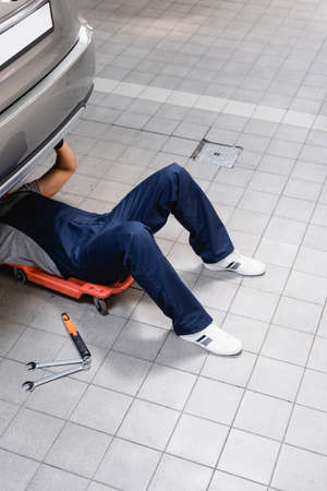 mechanic in uniform lying under car near metallic tools