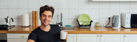 Panoramic shot of handsome man smiling at camera and holding cup of coffee in kitchenの写真素材