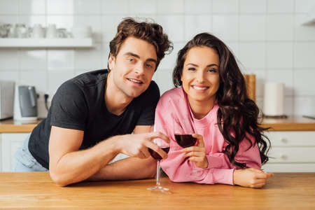 Young couple smiling at camera while toasting with wine at kitchen table