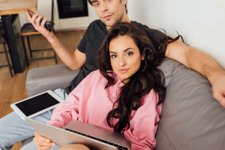 Selective focus of young couple looking at camera while using digital devices on couch