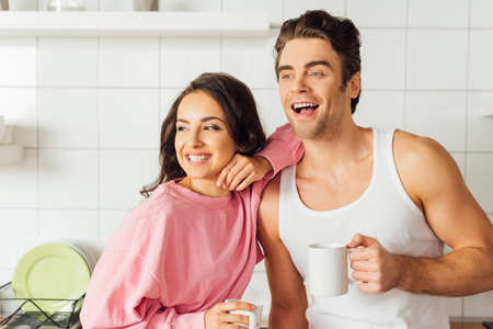 Positive woman holding cup of coffee and embracing handsome boyfriend in kitchenの写真素材
