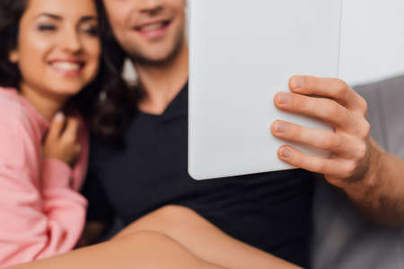 Selective focus of man holding digital tablet near cheerful girlfriend on couch isolated on gray